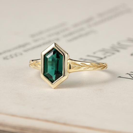 Hexagonal emerald ring in gold with leaf motifs, placed on an open book.