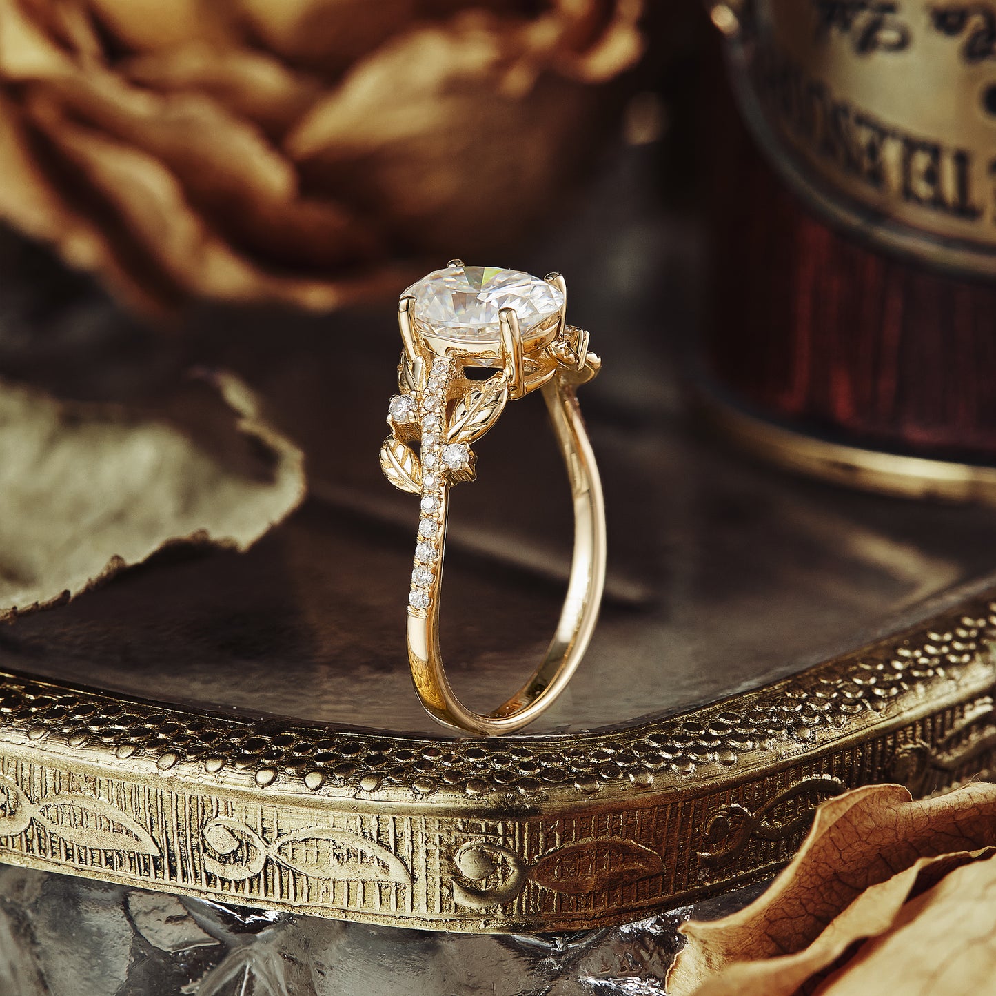 Moissanite engagement ring with oval cut, gold band, and leaf accents on a decorative tray.