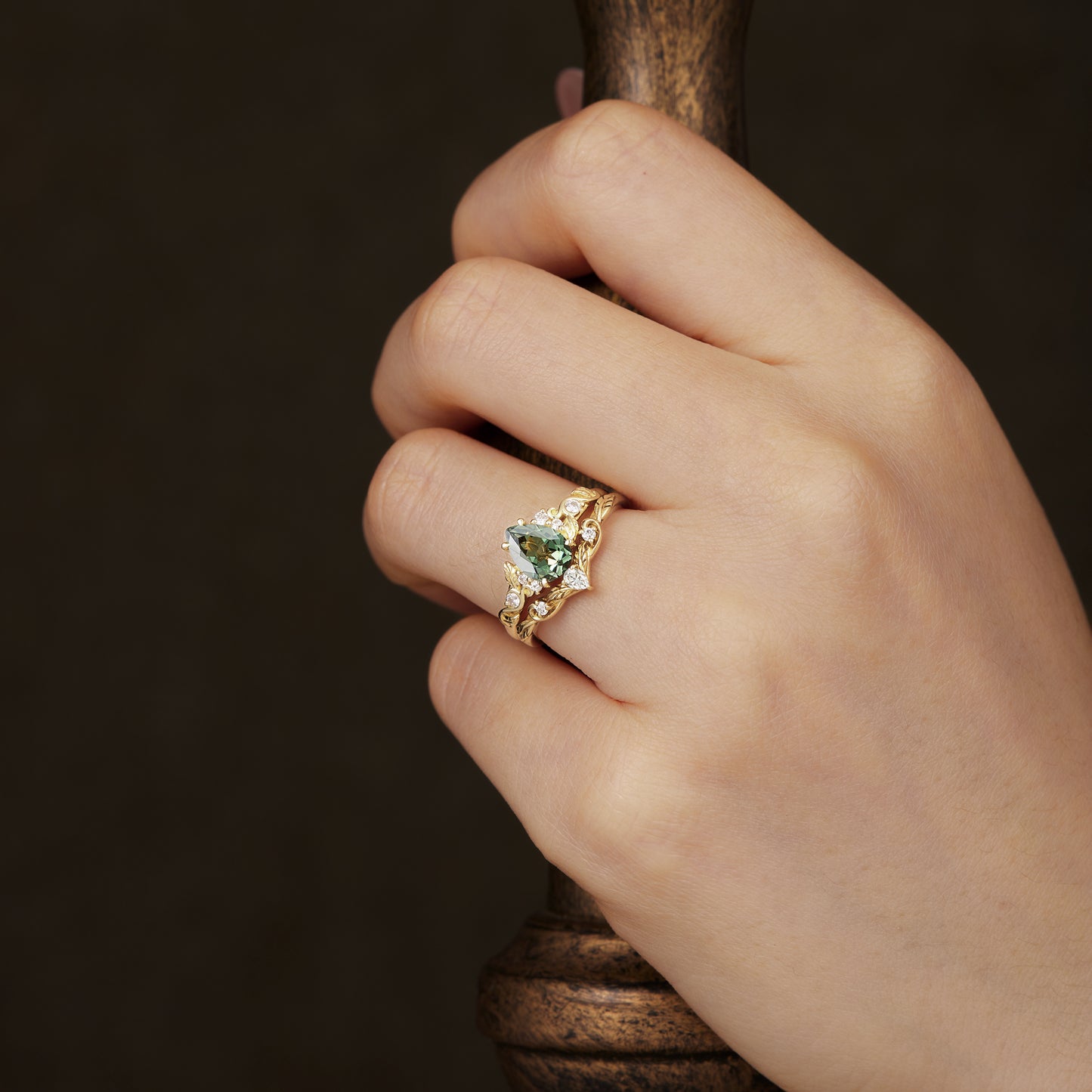Gold leaf ring set with pear-shaped green sapphire, worn on a finger.
