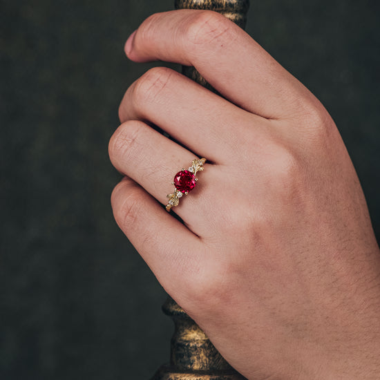 Hand displaying ruby engagement ring with moissanite side stones on gold band.