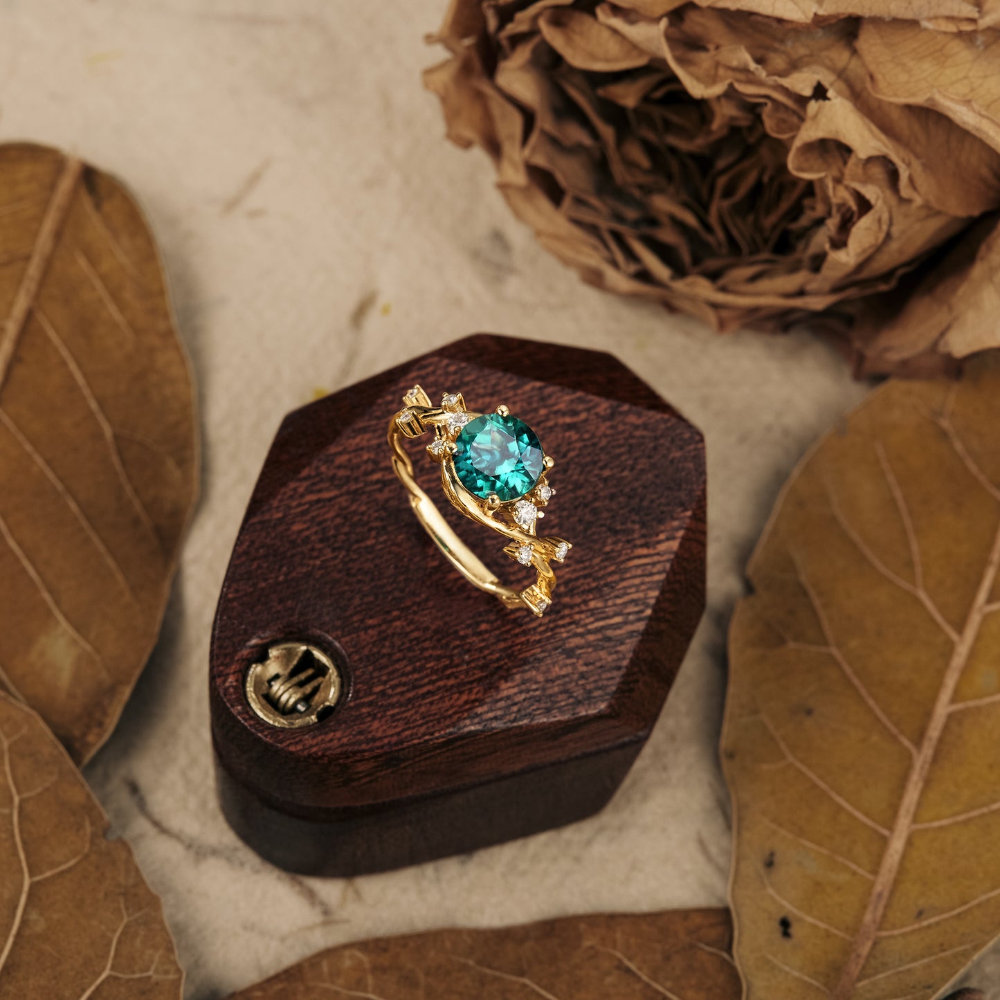 Teal round-cut gemstone ring on wooden box with gold entwined band and diamonds.