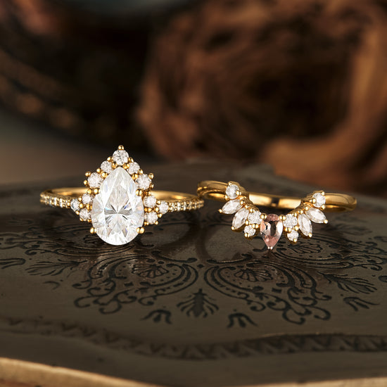 Gold pear Moissanite engagement ring with halo and floral wedding band set on decorative base.