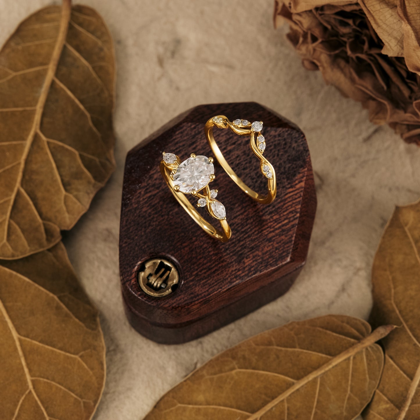 Moissanite engagement and wedding rings set on a wooden display with dried leaves.