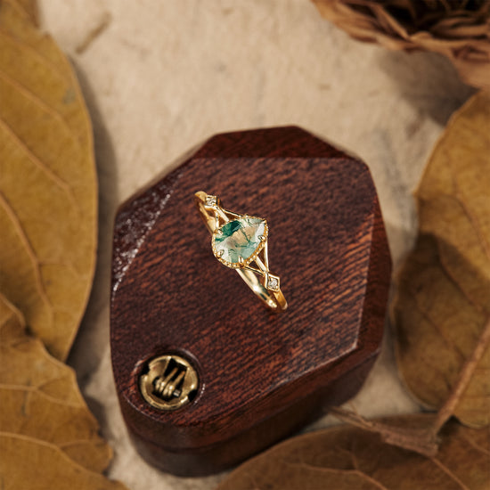 Nature-inspired moss agate pear ring on wooden box with leaf accents.