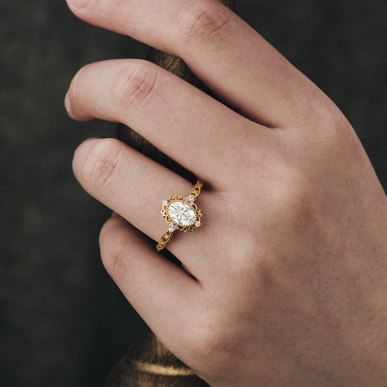 Ornate gold engagement ring featuring oval moissanite on a hand.